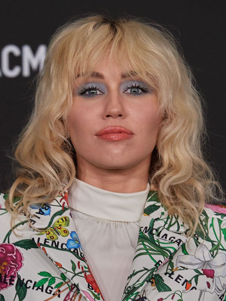 Mylie Cyrus at event with blonde softly wavy wolf cut hairstyle, wearing blue eyeshadow and orange lipstick