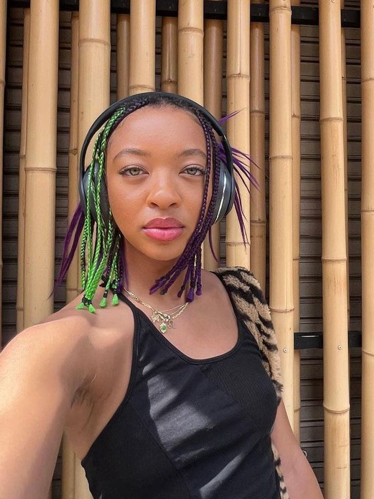 Woman with medium-length purple box braids styled with a few green braids, with loose spiked hair in back