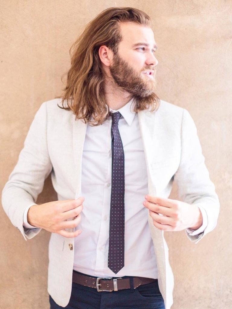 Viking Hairstyles: Man with rugged beard with shoulder length light brown wavy hair, wearing white top with black tie and white suit jacket outside