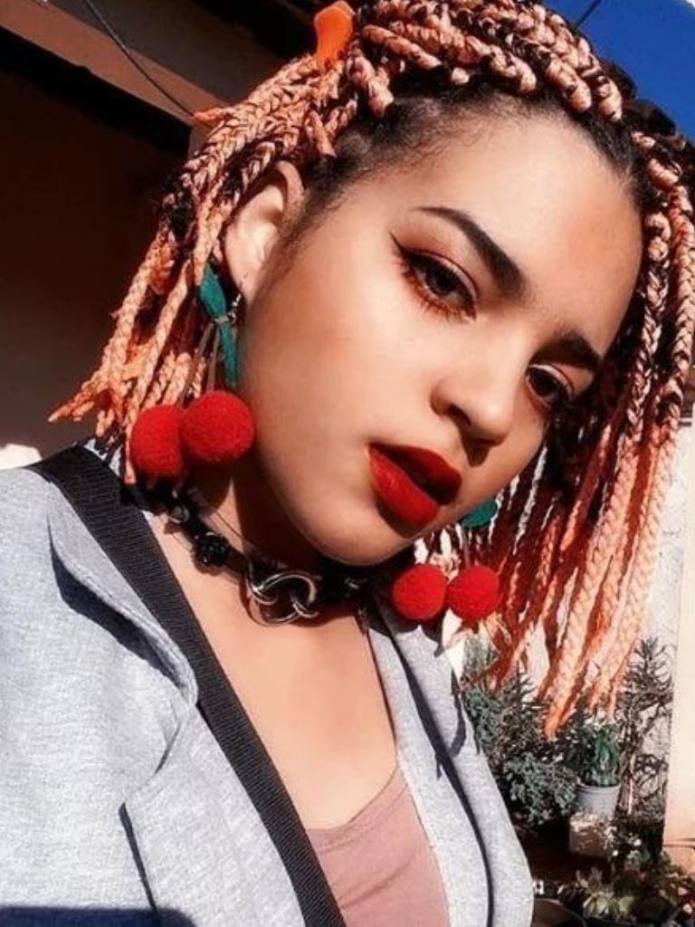 Bob braids bob: Woman with rose gold short box braids, wearing cherry earrings with grey blazer and pink top, posing outside