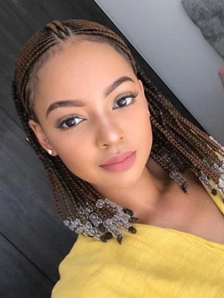 Bob braids bob: Woman with caramel brown short fulani braided bob hairstyle with bead detailing at the ends, wearing a yellow top and posing in a bathroom setting