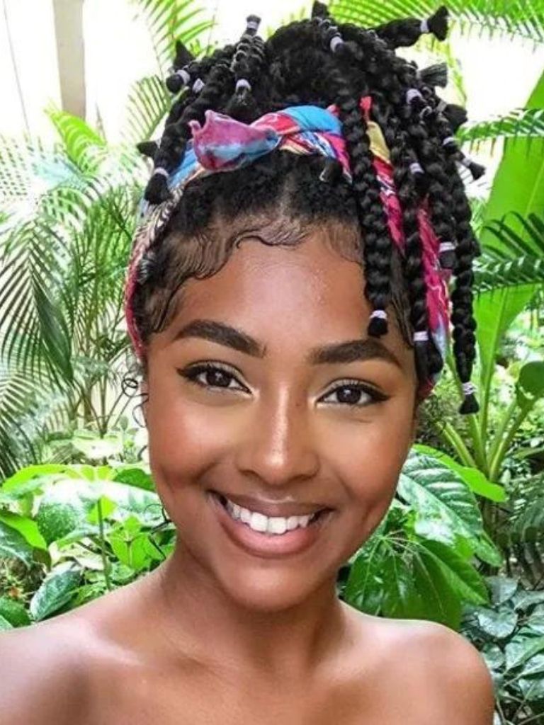 Box braids bob: Woman with dark brown box braids styled into a scarf updo, posing against some green plants