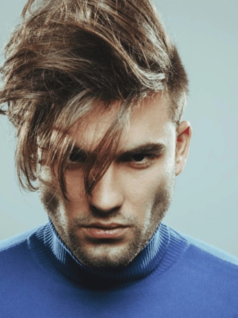 Men's haircuts for round faces: front facing image of a man with a fringe - mens haircuts for round faces: A man with medium brown hair with a long wavy fringe, wearing a blue jumper and posing in a studio setting