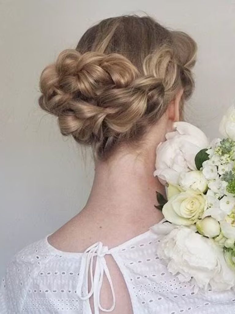 Updos with braids: Back shot of a woman with bronde hair styled into a low pull through braided bun.