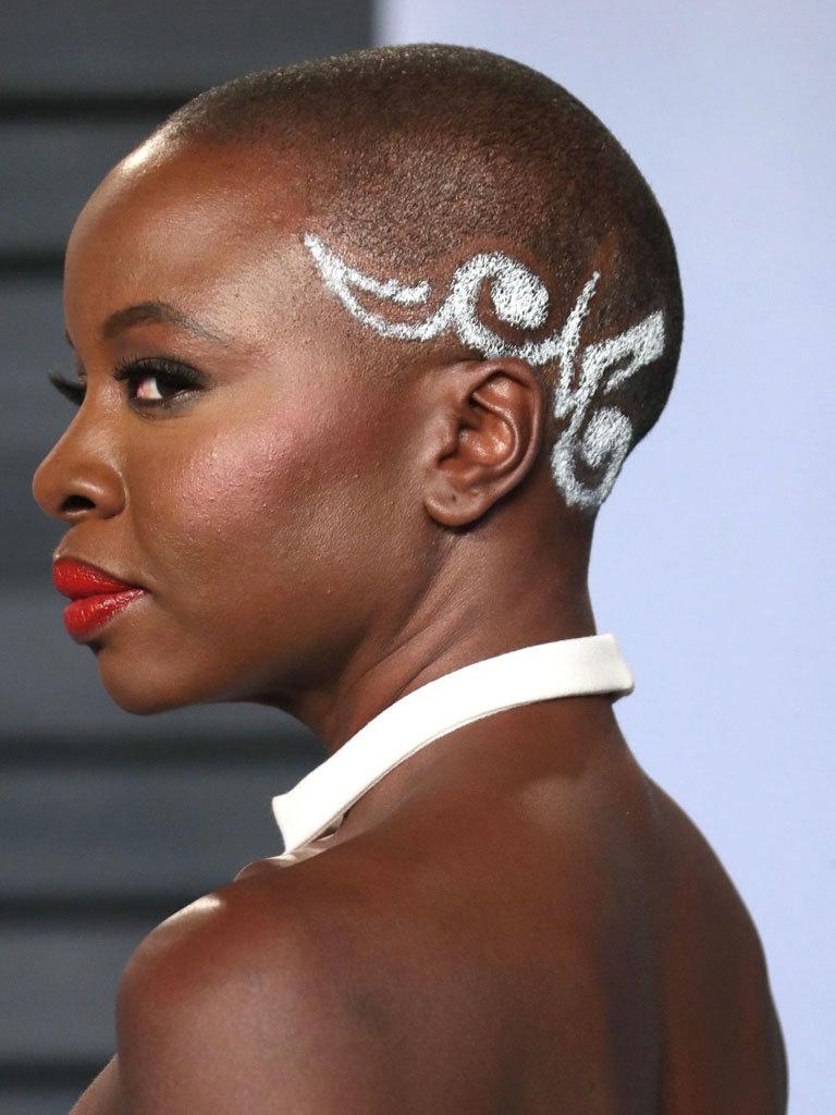 danai gurira with a shaved buzzcut with white pattern design on the side at movie awards wearing a white dress