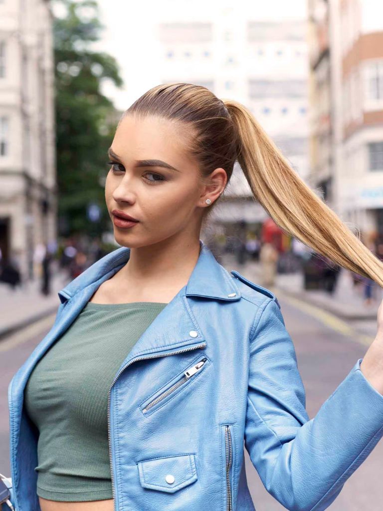 Club hairstyles: Image of a blonde woman with a sleek high ponytail wearing a blue leather jacket and khaki green top, standing in the middle of the road