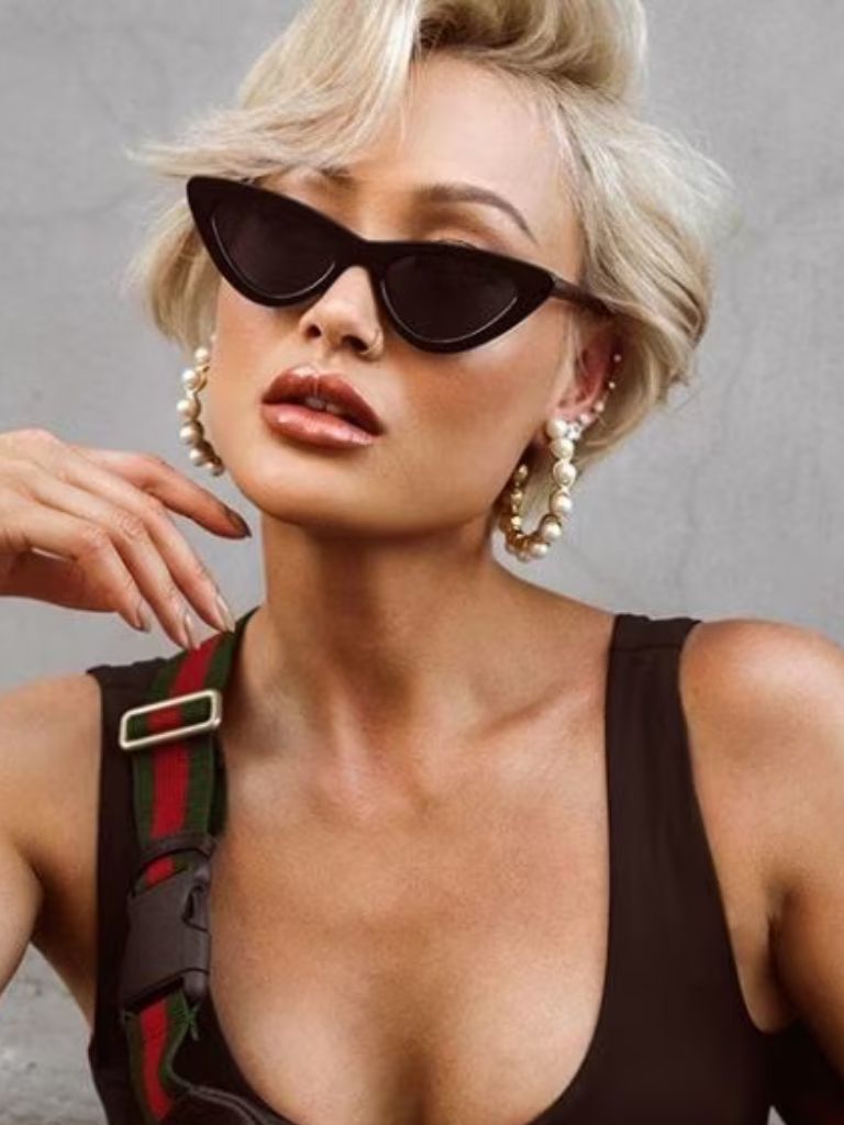 close up shot of woman with short pixie with side-swept bangs, wearing black top and posing with micro sunglasses