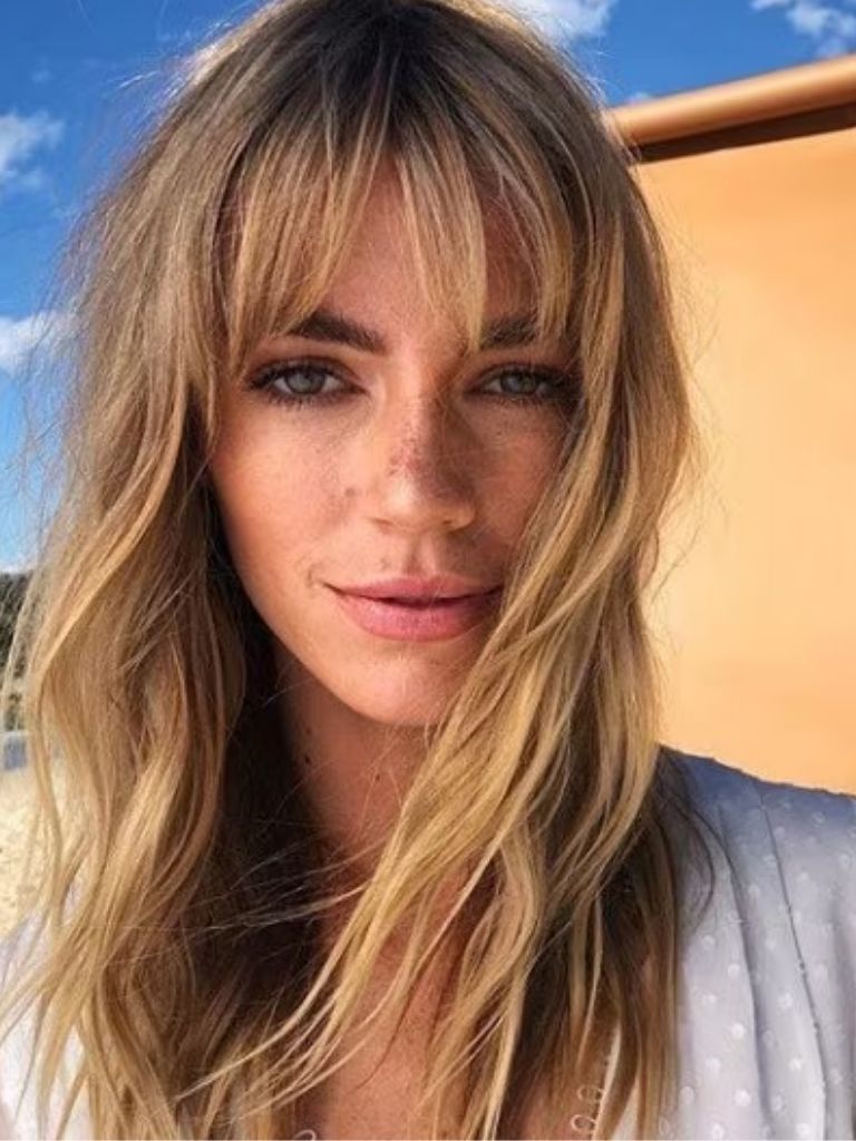 Types of bangs: Woman with long blonde hair with full Birkin bangs, wearing white and posing outside