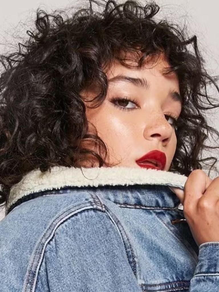 types of bangs: close up shot of woman with curly bangs hairstyle, wearing denim jacket and posing