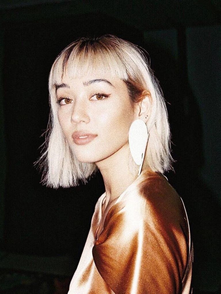 Types of bangs guide: Kim Jones with a bleach blonde long bob styled with a micro fringe, wearing a seashell earring with a satin orange top