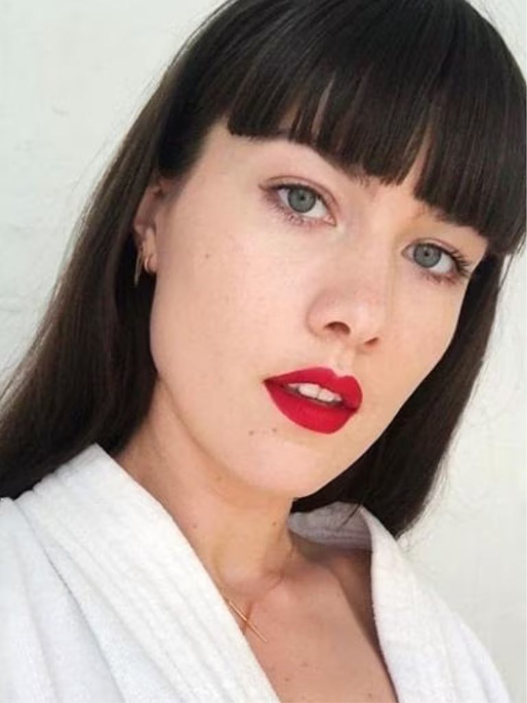 Type of bangs guide: Woman with long dark brown hair and blunt bangs, wearing white top, with white lipstick and posing against a white wall