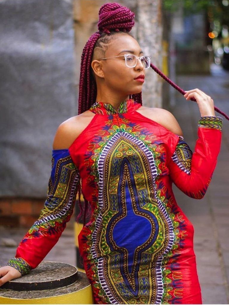Woman with shaved side hair and long red box braids styled into half-up bun wearing a red patterned dress.