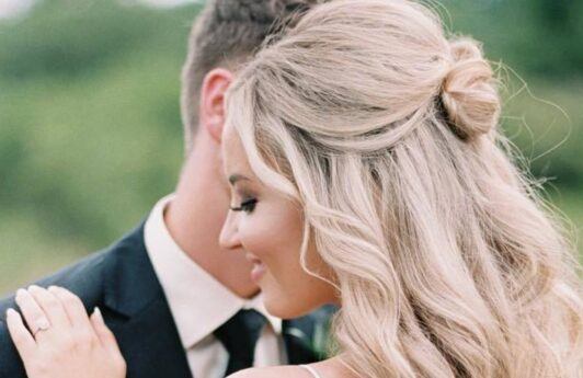 Half up half down wedding hairstyles: Woman with long bleach blonde curly styled into a half-up bun, posing outside.