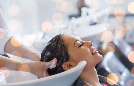 beauty, hair care and people concept - happy young woman with hairdresser washing head at hair salon over holidays lights