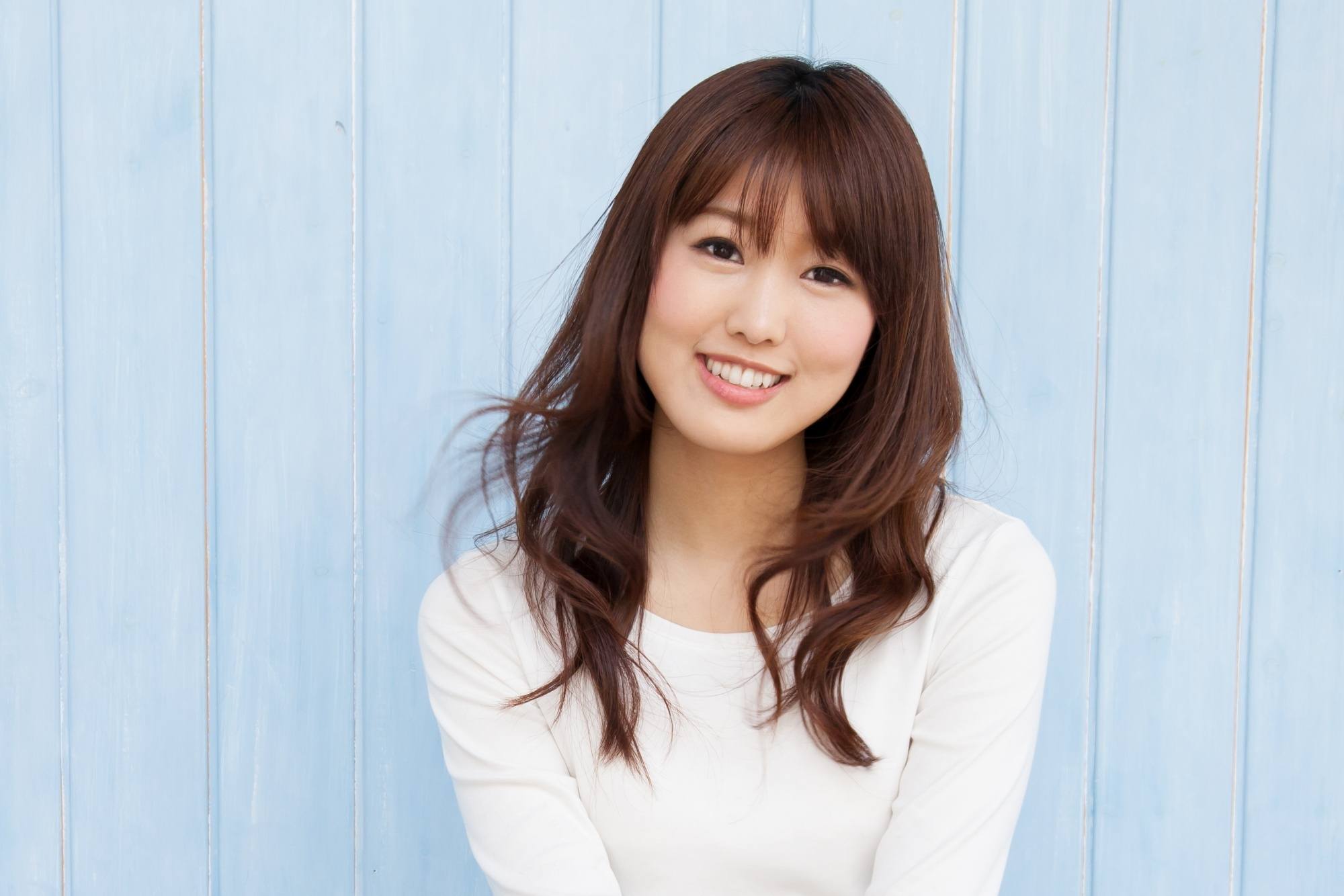 Asian woman with long wavy hair and wispy bangs wearing a white top.
