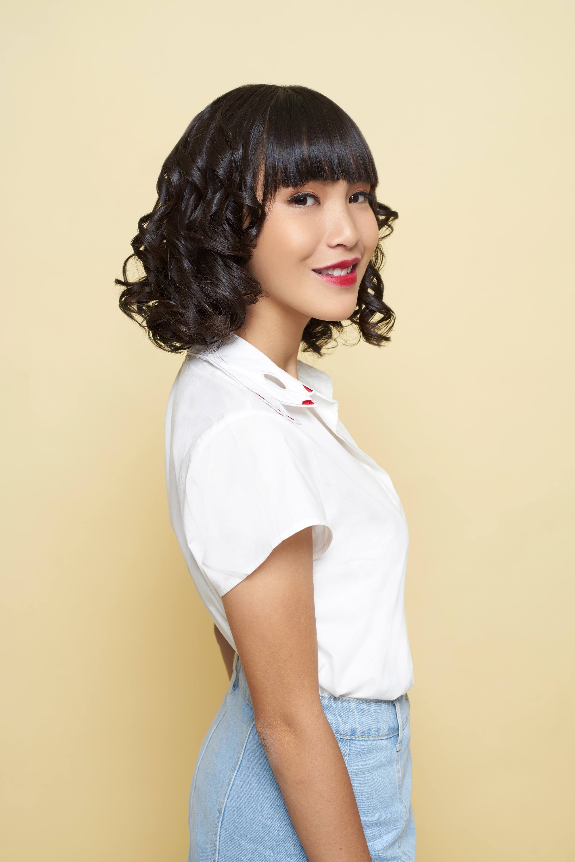 Asian woman with short curly hair with bangs wearing a white top and denim skirt smiling