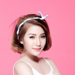 Chin length hairstyles: Closeup shot of Asian woman wearing white thin-strapped top with brown short hair and white headband standing against pink background