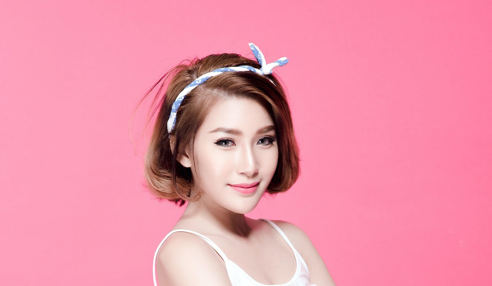 Chin length hairstyles: Closeup shot of Asian woman wearing white thin-strapped top with brown short hair and white headband standing against pink background