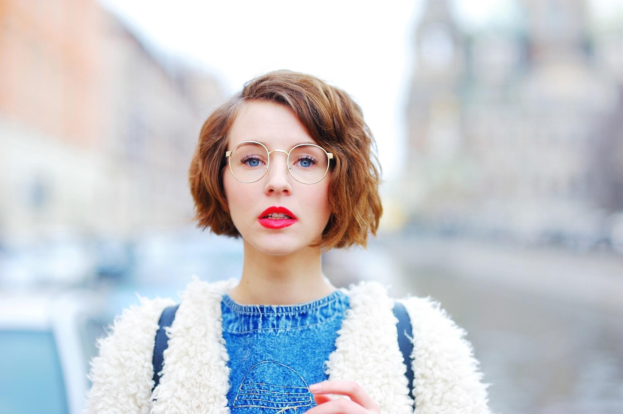 Chin length hairstyles: Closeup shot in outdoor location of a woman wearing white jacket and blue shirt with brown wavy bob and eye glasses