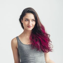 Dip dyed hair: Woman wearing a gray tank top with long curly dip dyed magenta hair standing against a white background