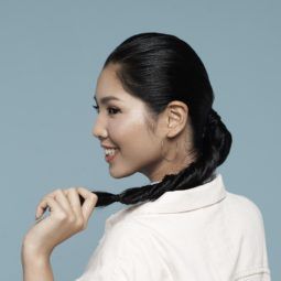 Side view of Asian woman wearing long-sleeved polo shirt standing against blue background with long black hair in double rope braid ponytail