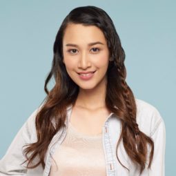 Face framing lace braid: Asian woman with long brown hair in lace braid wearing light peach blouse, white blazer, and pink skirt standing against a blue background with right and on hips