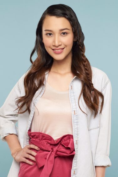 Face framing lace braid: Asian woman with long brown hair in lace braid wearing light peach blouse, white blazer, and pink skirt standing against a blue background with right and on hips