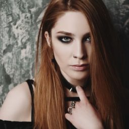 Goth hairstyles: Closeup shot against a stone background of a woman wearing a black top, dark eyeliner, and has long brown hair partially covering the face