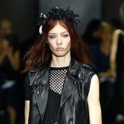 Goth hairstyles: Runway shot oman wearing black leather vest and black see-through top with reddish brown hair in a textured hairstyle topped with a black hair pece