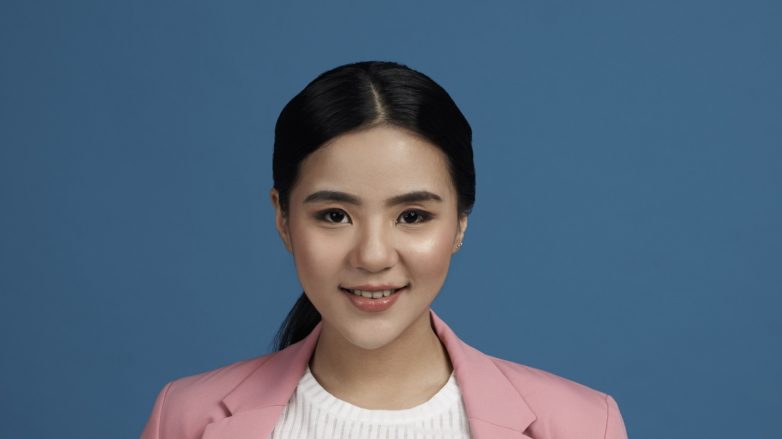 Tucked in ponytail: Asian woman wearing white blouse and pink blazer with long black hair in ponytail standing against a blue background