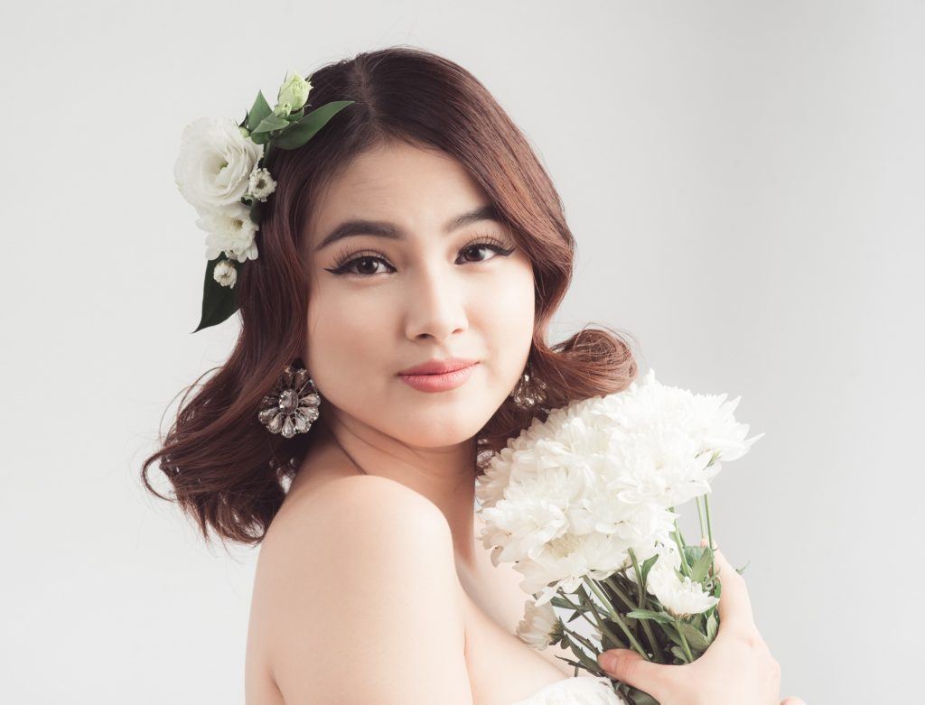 Wedding hairstyles for medium hair: Closeup shot of a bride with shoulder-length wavy brown hair with floral hair piece holding a white bouquet standing against a light gray background