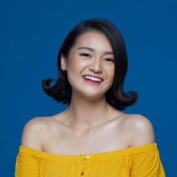Flipped-out short bob: Closeup shot of an Asian woman with short black hair wearing a yellow dress against a blue background