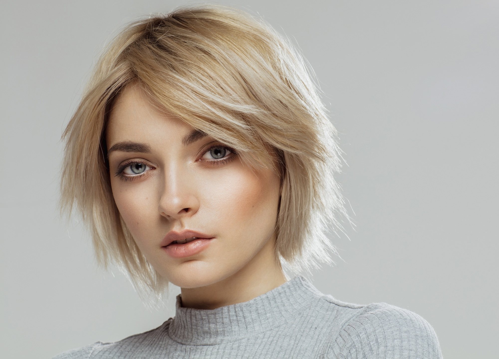 Haircuts for rainy season: Closeup shot of a woman with short layered blonde hair wearing a gray top against a gray background