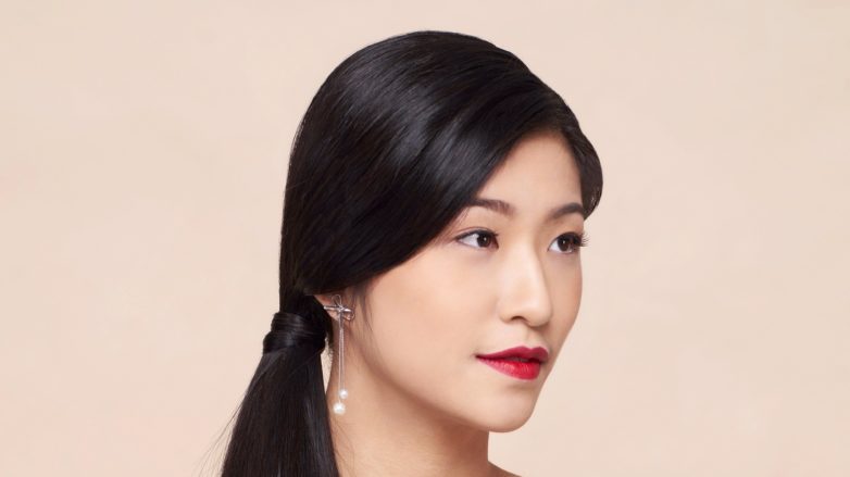 Side ponytail: Asian woman wearing dark blue blouse with long black hair in side ponytail against an oyster-colored background