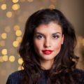 Christmas hairstyles: Closeup shot of a woman with dark wavy hair wearing dark blue blouse with Christmas lights at the background