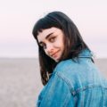 Should I get bangs: Woman with black hair and baby bangs wearing a denim jacket by the beach