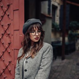 Should I get bangs: Woman with long wavy hair and wispy bangs with eyeglasses wearing a coat and bonnet beside a red gate