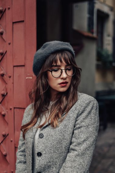 Should I get bangs: Woman with long wavy hair and wispy bangs with eyeglasses wearing a coat and bonnet beside a red gate