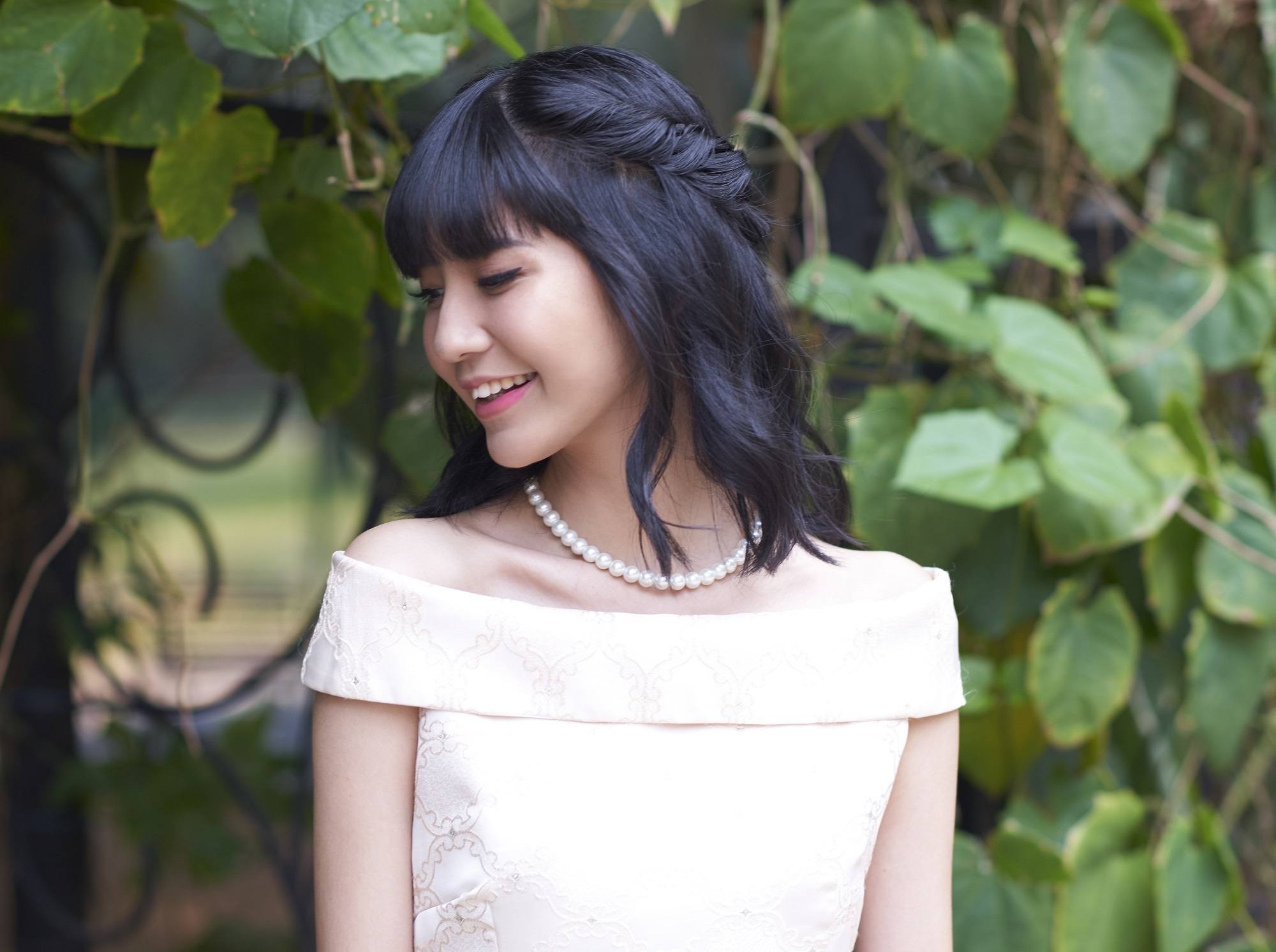 Valentine hairstyles: Asian woman wearing a peach dress with shoulder length hair in a half updo standing against a leafy gate