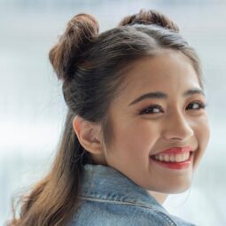 Asian woman with space buns for short hair smiling and wearing a denim jacket.