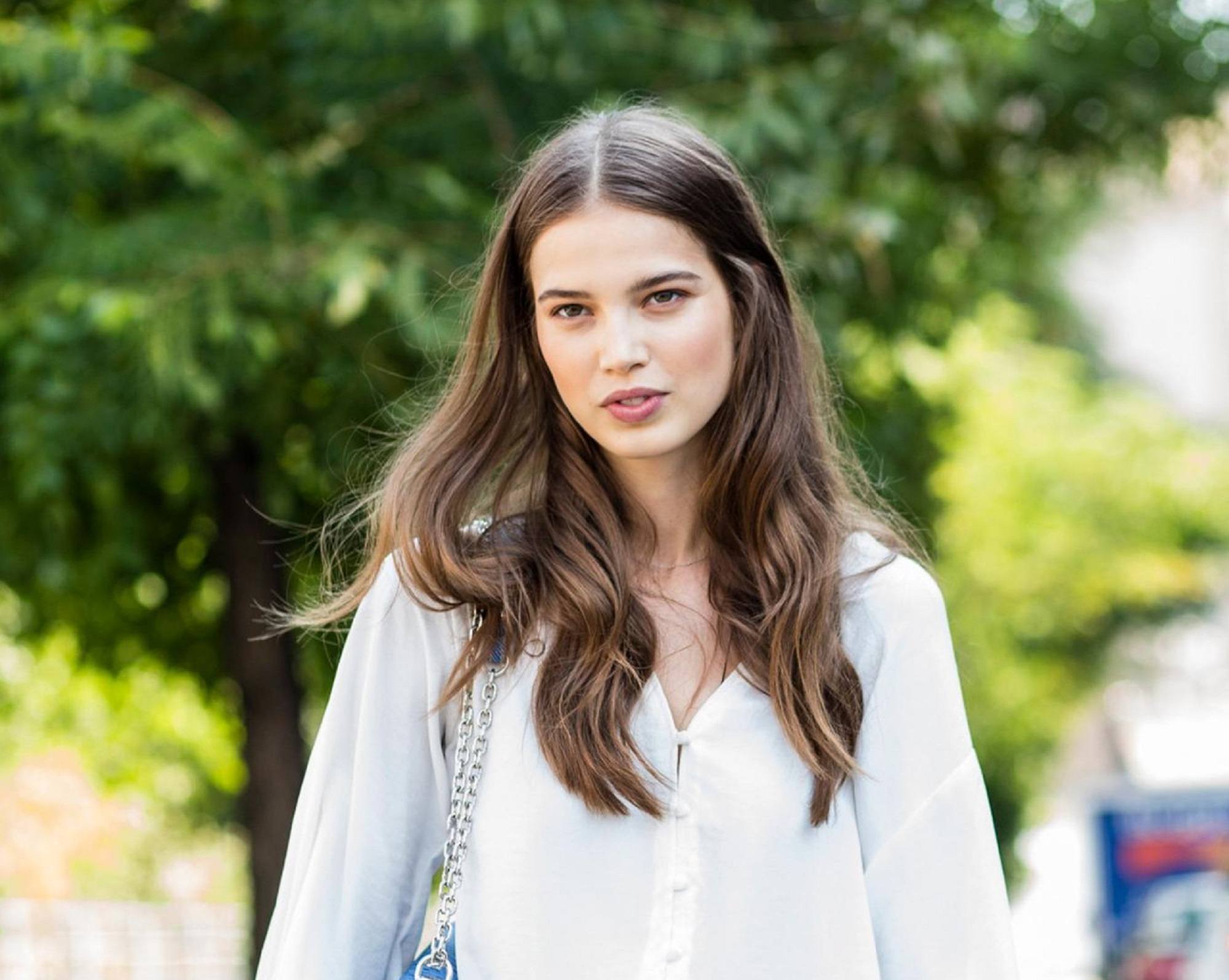 Woman with long wavy ash brown hair wearing a white blouse standing outdoors