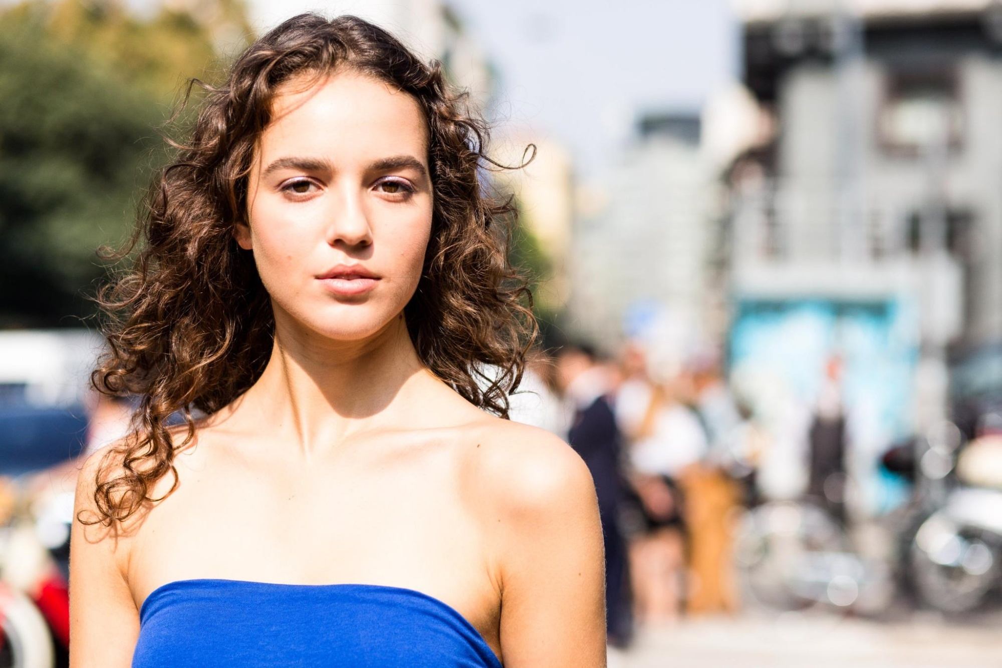 Caucasian woman with long brown curly hair wearing a blue tube top outdoors