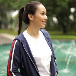 Asian woman with long hair in a fishtail braid ponytail wearing a jacket and smiling in front of a fountain