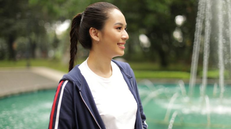 Asian woman with long hair in a fishtail braid ponytail wearing a jacket and smiling in front of a fountain