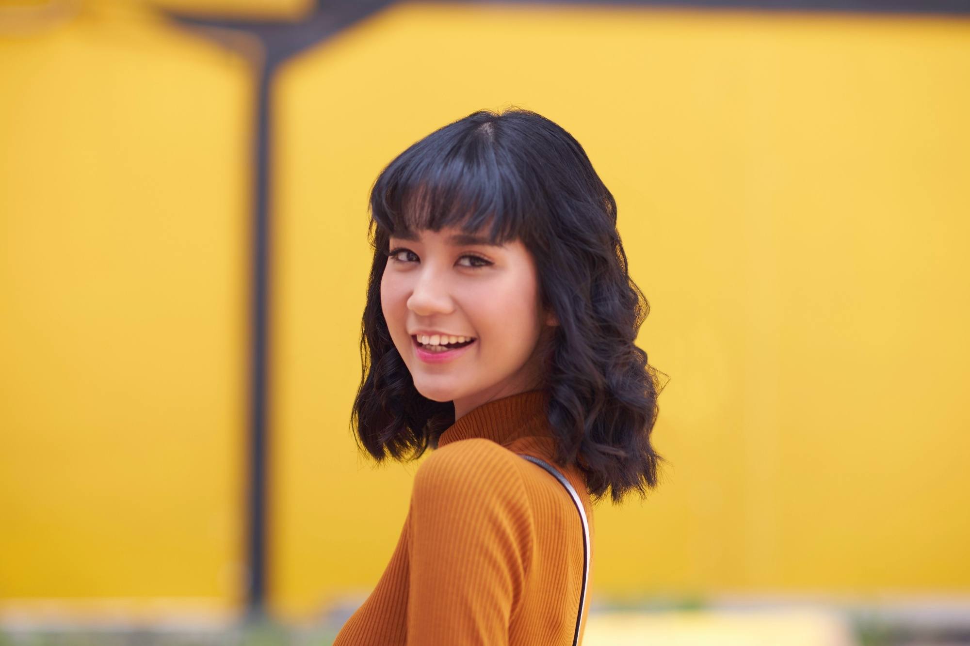 Asian woman with curly short hair with bangs wearing a mustard top