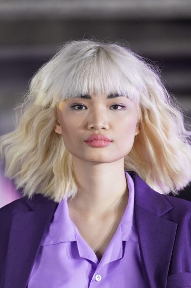 Asian woman with wavy blonde hair with bangs wearing a purple outfit