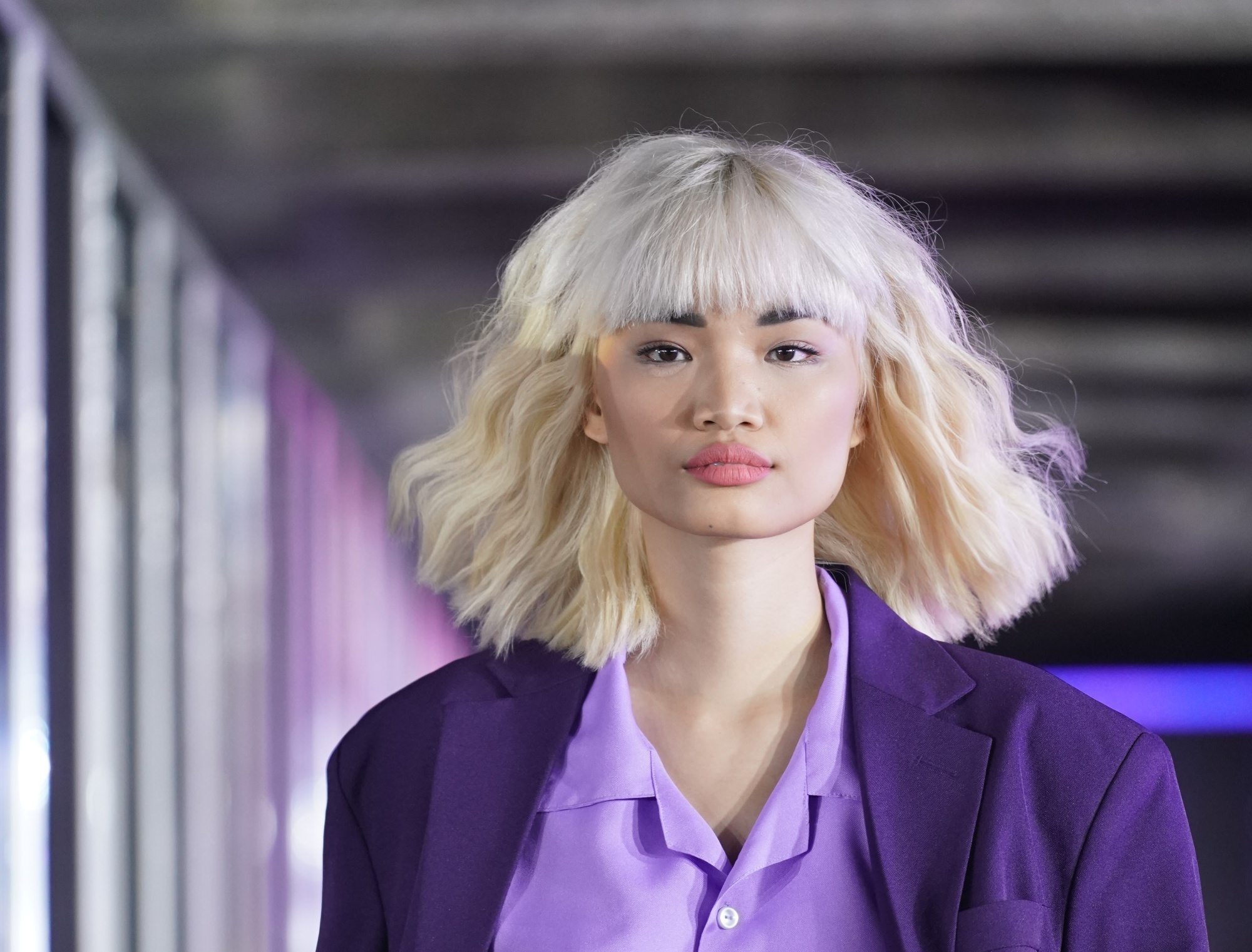 Asian woman with wavy blonde hair with bangs wearing a purple outfit