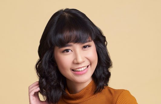 Asian woman with wavy lob and blunt bangs wearing a brown top and black skirt