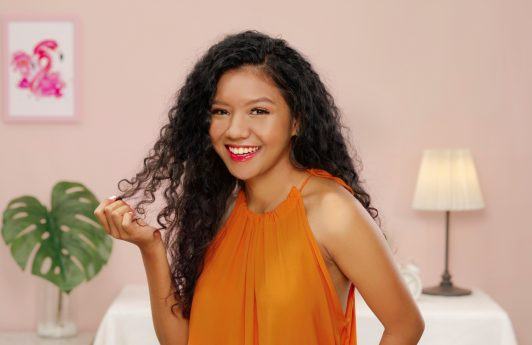 Asian woman with long curly hair smiling and wearing an orange sleeveless top