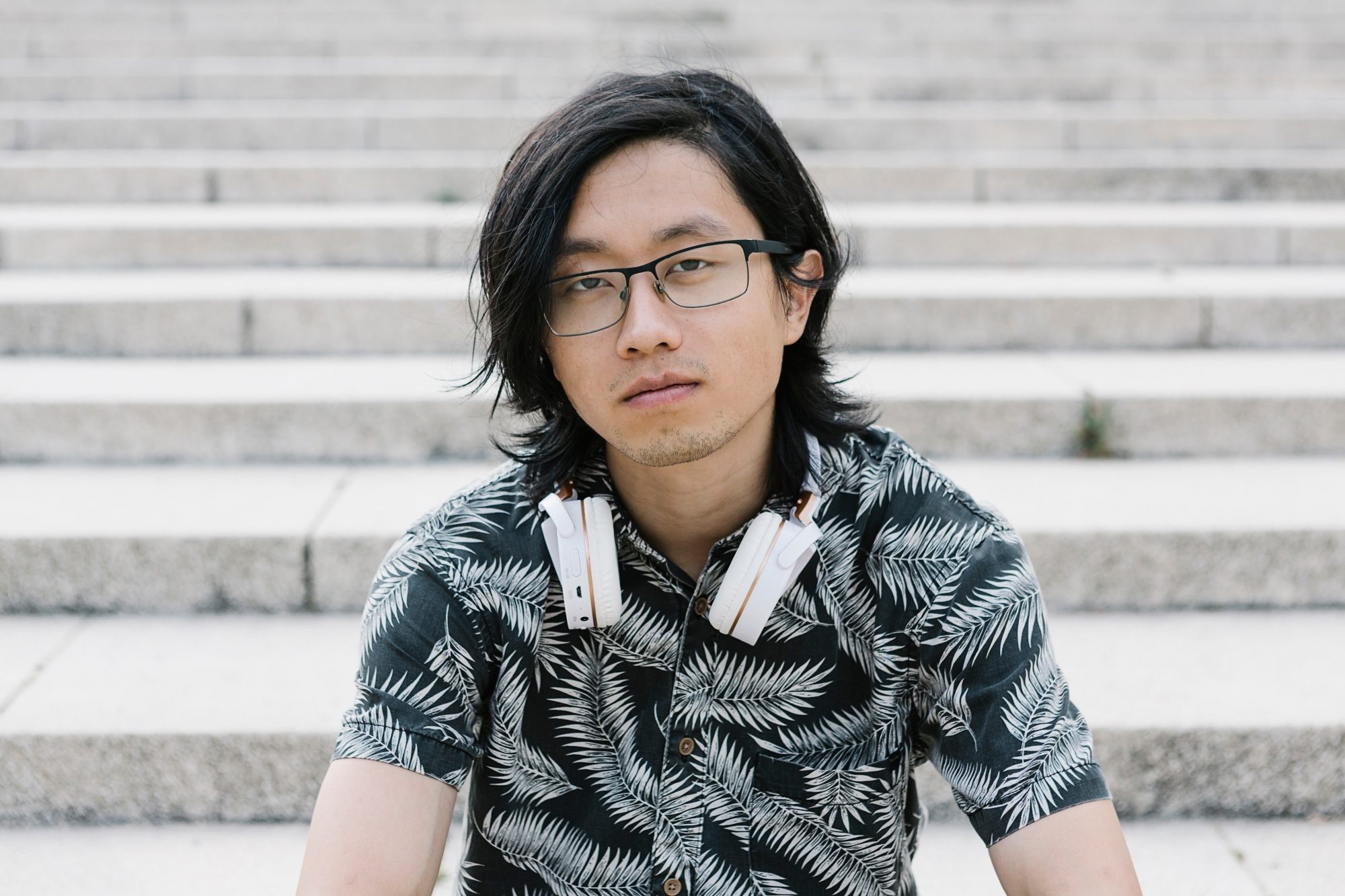 Asian man with long hair wearing eyeglasses sitting on outdoor steps
